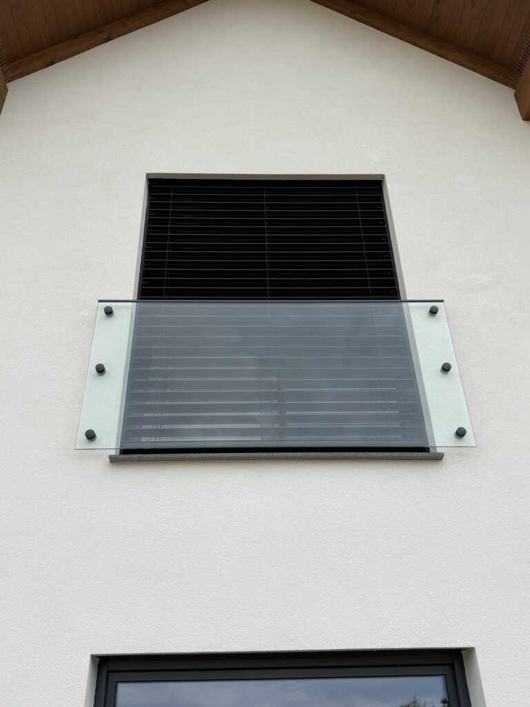 Balkon francuski z tafli szkła mocowanej punktowo do elewacji przed oknem z żaluzją