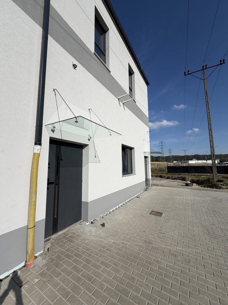 Daszek ze szkła na nierdzewnych cięgnach nad drzwiami, widok wzdłuż elewacji budynku