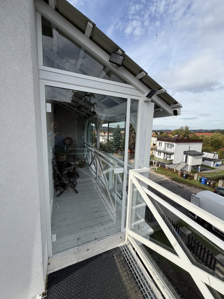 Balkon na piętrze zabudowany szklanymi taflami w białej konstrukcji, z przeszklonym szczytem