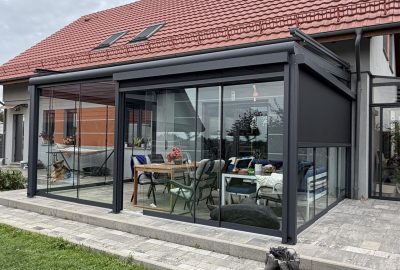Taras zabudowany szklanymi ścianami przesuwnymi w grafitowej konstrukcji przy domu z czerwoną dachówką